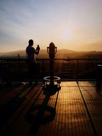 Silhouette people sitting by railing against sky during sunset