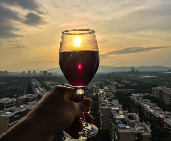 Close-up of hand holding drink against cityscape during sunset