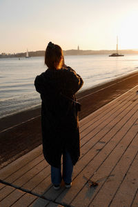 Rear view of woman standing at beach against sky during sunset