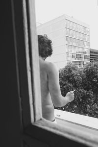 Shirtless man with cigarette standing in balcony seen through window