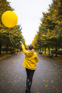 Rear view of woman with yellow helium balloon walking on road at park during autumn