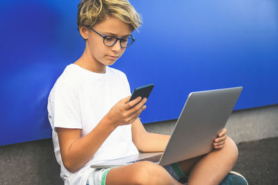 Boy using technologies while sitting against blue wall