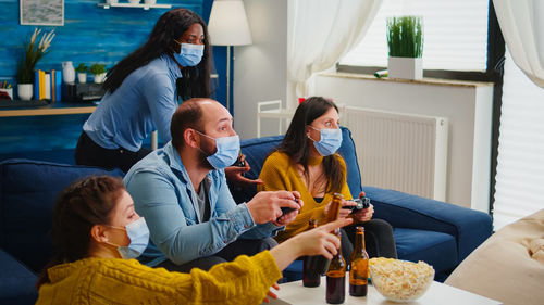 Cheerful people wearing mask playing video game while sitting on sofa at home