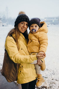 Portrait of smiling mother carrying son o footpath during winter