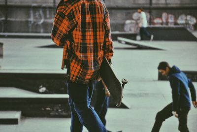 Man walking with skateboard in skateboard park with friends skating in background