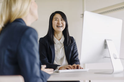 Two laughing businesswomen sitting at desk in office talking