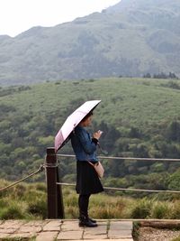 Side view of young woman with umbrella standing against mountains