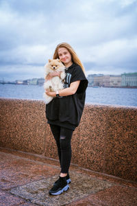 Cheerful and happy girl teenager holds and hugs pomeranian spitz on embankment