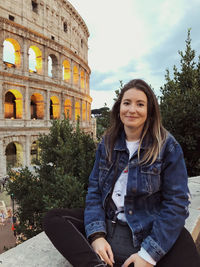 Portrait of young woman sitting on railing against coliseum