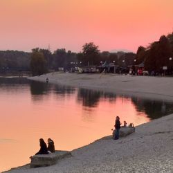 People sitting by lake against orange sky