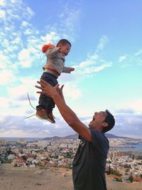 Side view of playful father throwing son in air against cityscape and cloudy sky