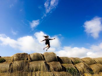 Low angle view of excited woman jumping over hay bales on field against sky