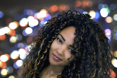 Close-up portrait of young woman with curly hair against illuminated lights