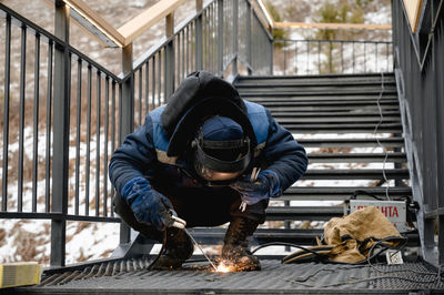 A man wearing work clothes will weld a ladder in the mountains. the welder is wearing a blue uniform 