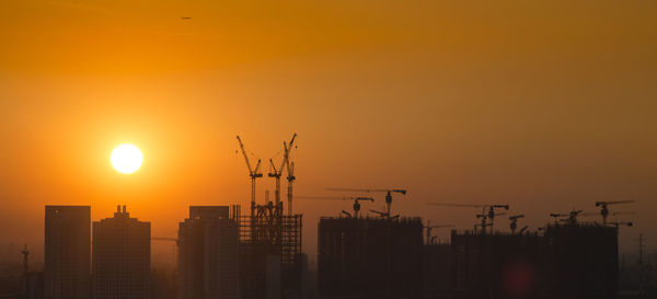 Silhouette cranes by buildings against sky during sunset