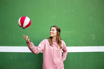 Smiling young woman tossing basketball while standing against wall