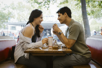 Smiling man holding burger and glancing at girlfriend sitting in restaurant