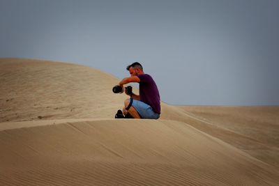 Man holding camera while sitting on sand dune