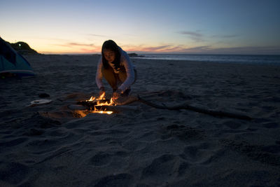 Woman standing by campfire on beach against sky during sunset