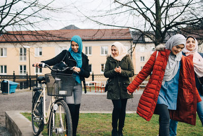 Happy multi-ethnic female friends walking with bicycle on grass in city