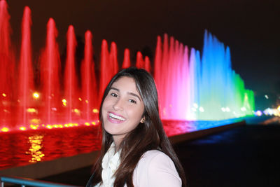 Portrait of smiling woman by illuminated fountain at park during night