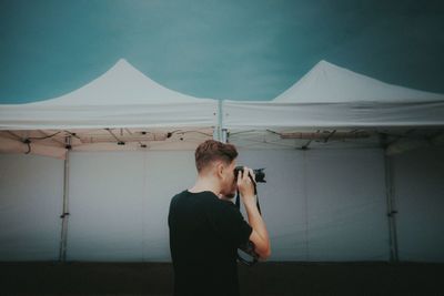 Rear view of young man photographing while standing by tent against sky