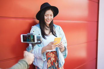 Happy young woman using mobile phone by cropped hand photographing against wall