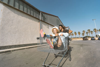 Young man pushing girlfriend sitting in shopping cart outside supermarket