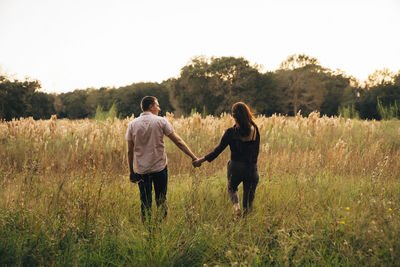 Rear view of couple holding hands while walking on grassy field against clear sky at park