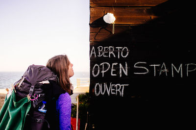 Female hiker looking at sign on wooden wall against sky