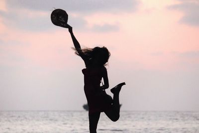 Silhouette woman holding hat while jumping at beach against sky during sunset