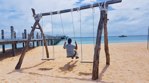 Rear view of man on swing at beach against cloudy sky