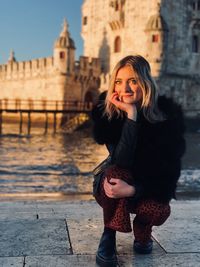 Portrait of smiling young woman crouching by river against building in city