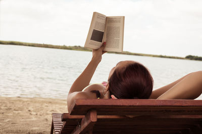 Woman reading book while lying on bench at beach against cloudy sky