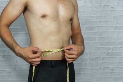 Midsection of shirtless man measuring waist with tape measure by wall