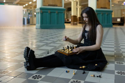 Beautiful girl in black dress plays chess in hall checkered floor