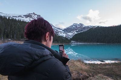 Rear view of man photographing lake against snowcapped mountains