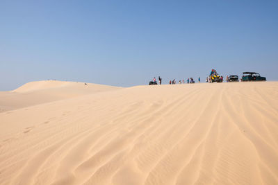 Low angle view of people with vehicles on sand dune in desert against blue sky