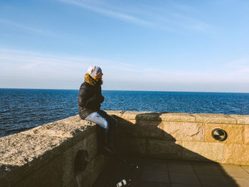 Man sitting on retaining wall by sea against sky