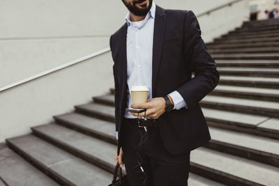 Midsection of businessman with disposable cup and smart phone standing on staircase