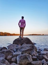 Rear view of man standing on rocks by sea against sky during sunset