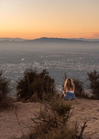 Rear view of woman sitting against cityscape against sky during sunset