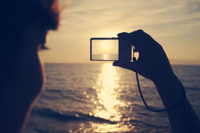 Close-up of woman photographing sea with digital camera against sky during sunset