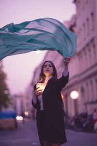 Portrait of woman moving green scarf while holding illuminated jar in city at dusk