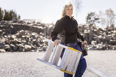 Smiling female electrician carrying ladder while walking on road