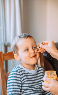 Mom feeds a little boy with a spoon. the boy is sitting on a chair at the table