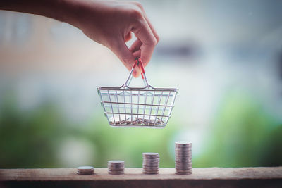 Cropped hand of person holding small shopping basket by stacked coins outdoors