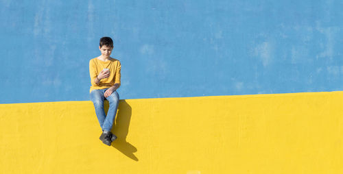 Boy using phone while sitting on yellow railing against blue wall