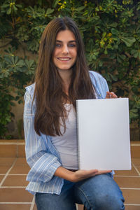 Portrait of smiling woman holding spiral notebook against plants