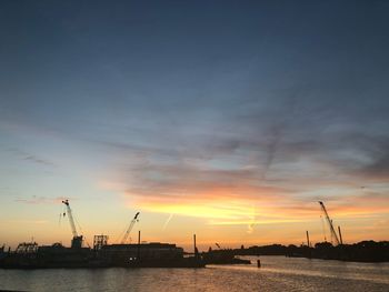 Silhouette cranes at harbor against sky during sunset